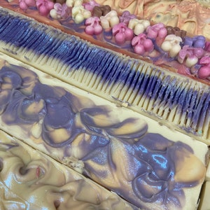 May include: Close-up of several bars of handmade soap. The soaps feature various designs, including floral embellishments and swirled patterns in shades of purple, pink, white, and brown. The soaps are rectangular and appear to be freshly cut.