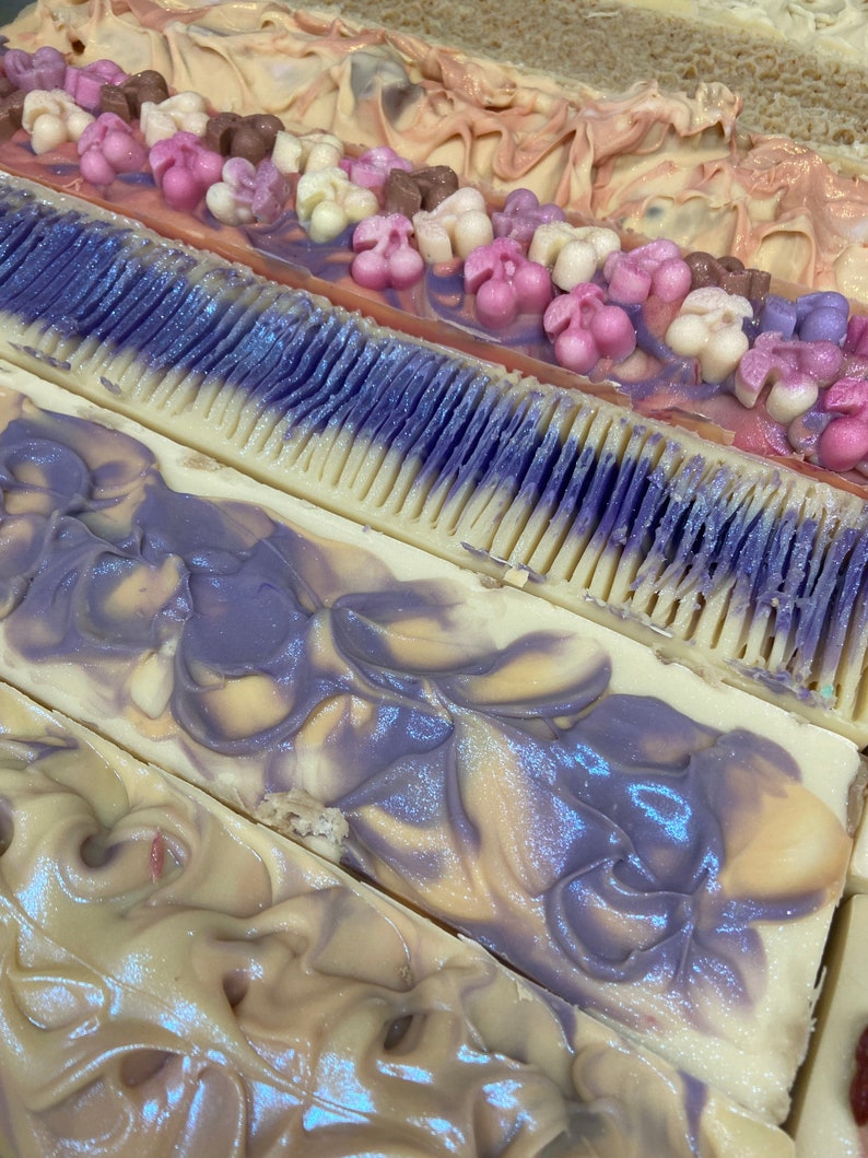 May include: Close-up of several bars of handmade soap. The soaps feature various designs, including floral embellishments and swirled patterns in shades of purple, pink, white, and brown. The soaps are rectangular and appear to be freshly cut.