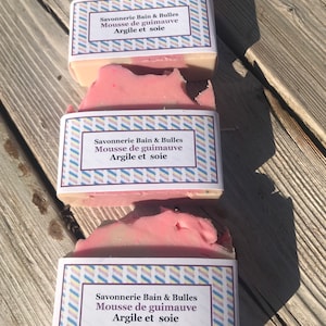 May include: Three handmade soaps with pink and white layers are stacked on a wooden surface. Each soap has a label with French text: "Savonnerie Bain & Bulles, Mousse de guimauve, Argile et soie."