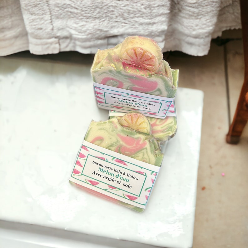 Op de afbeelding: Twee handgemaakte zeepjes met een watermeloen- en limoensmaak. De zeep is verpakt in roze en groen papier met de tekst "Savonnerie Bain & Bulles Melon d'eau Avec argile et soie" erop gedrukt.