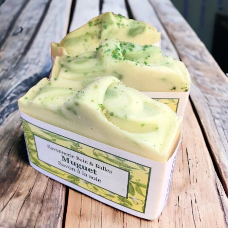 May include: Close-up of three bars of handmade soap. The soap is a creamy white color with green swirls and speckles. Each bar is labeled with the text "Savonnerie Bain & Bulles Muguet Savon &agrave; la soie". The soap is displayed on a wooden surface.