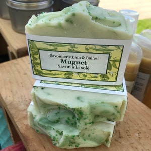 May include: Stacked bars of handmade soap with a light green hue and speckled green accents. Each bar is wrapped with a label that reads "Savonnerie Bain & Bulles Muguet Savon &agrave; la soie."