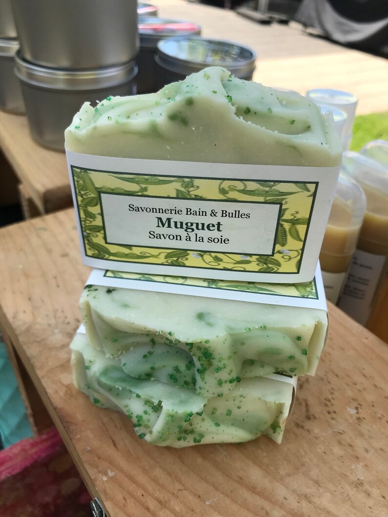 May include: Stacked bars of handmade soap with a light green hue and speckled green accents. Each bar is wrapped with a label that reads "Savonnerie Bain & Bulles Muguet Savon &agrave; la soie."