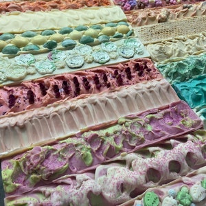 May include: A collection of handmade soap bars in various colours and designs. The bars are arranged in rows, with each row featuring a different pattern or colour. Some of the bars have glitter, while others have swirls or flowers.