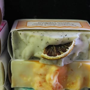 Puede incluir: Primer plano de barras de jabón hechas a mano. Una barra es de color verde claro con motas oscuras y una rodaja de naranja seca. El jabón está envuelto en papel con texto. Se ven otras barras, con tonos amarillos y naranjas.