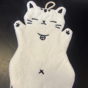 Op de afbeelding: Witte katvormige washand met zwarte accenten. De washand heeft een lachend kattengezicht met gesloten ogen, snorharen en een kleine tong. De kat heeft zwarte pootafdrukken en een lus om op te hangen.
