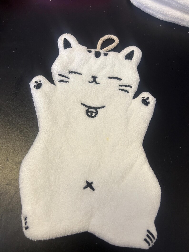 Op de afbeelding: Witte katvormige washand met zwarte accenten. De washand heeft een lachend kattengezicht met gesloten ogen, snorharen en een kleine tong. De kat heeft zwarte pootafdrukken en een lus om op te hangen.