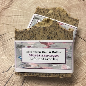May include: Two rectangular bars of handmade soap with a speckled, light brown appearance. Each bar is wrapped with a label that reads "Savonnerie Bain & Bulles Mures sauvages Exfoliant avec thé."