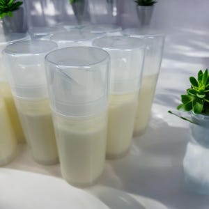 May include: A collection of cylindrical cosmetic pump bottles filled with an opaque off-white cream. Each bottle features a clear plastic cap and a white pump mechanism, arranged in a group to display the uniform packaging and contents.
