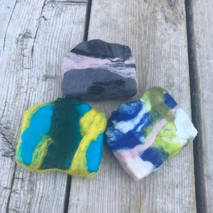 May include: Three felted soap bars in different colors and patterns. The soap bars are made of wool and have a soft, textured surface. The colors are blue, green, yellow, pink, and black.