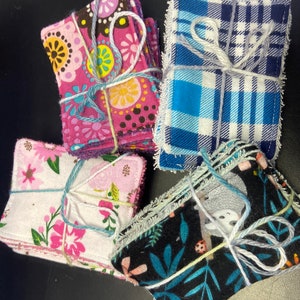 Op de afbeelding: Vier stapels herbruikbare make-up remover pads in verschillende stoffenpatronen. De pads zijn vastgebonden met touw en hebben bloemen-, ruit- en dierenprintontwerpen.