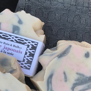 Puede incluir: Dos pastillas de jab&oacute;n de color blanco roto con remolinos de color rosa y gris oscuro. Una pastilla tiene una etiqueta de papel blanco con bordes geom&eacute;tricos negros y el texto Savonnerie Bain & Bulles Cerisier japonais Savon &agrave; la soie.