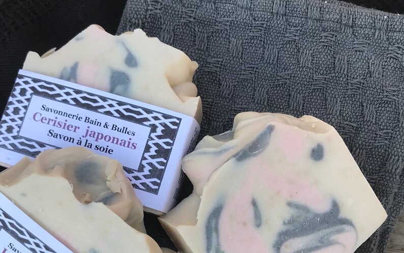 Puede incluir: Dos pastillas de jab&oacute;n de color blanco roto con remolinos de color rosa y gris oscuro. Una pastilla tiene una etiqueta de papel blanco con bordes geom&eacute;tricos negros y el texto Savonnerie Bain & Bulles Cerisier japonais Savon &agrave; la soie.