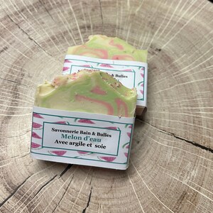 Op de afbeelding: Twee handgemaakte zeepjes verpakt in papier met watermeloenmotief. De zeep is groen en wit met roze swirls. Het etiket luidt "Savonnerie Bain & Bulles Melon d'eau Avec argile et soie".