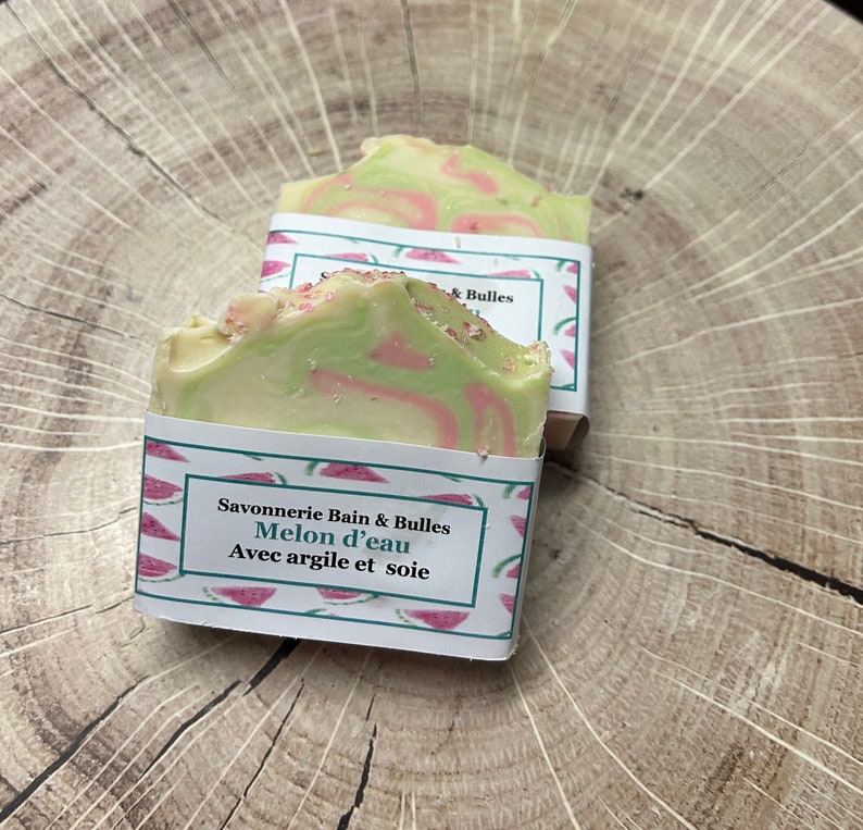 Op de afbeelding: Twee handgemaakte zeepjes verpakt in papier met watermeloenmotief. De zeep is groen en wit met roze swirls. Het etiket luidt "Savonnerie Bain & Bulles Melon d'eau Avec argile et soie".