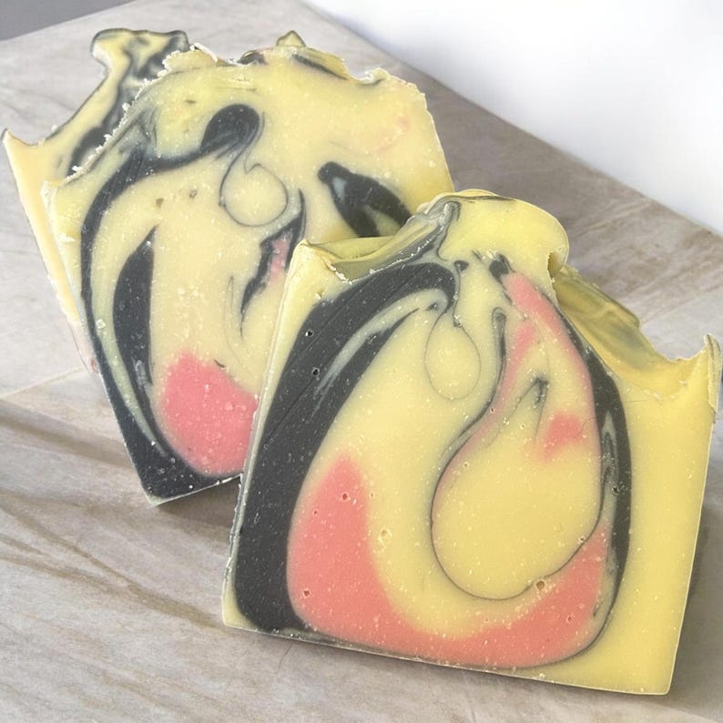 Puede incluir: Dos barras rectangulares de jab&oacute;n artesanal con un patr&oacute;n de remolinos marmolados. El jab&oacute;n presenta una paleta de colores amarillo p&aacute;lido, negro carb&oacute;n intenso y rosa coral suave, con una textura lisa y transiciones de color curvas.