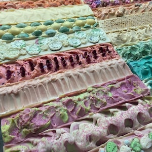 May include: A collection of handmade soap bars in various colours and designs. The bars are arranged in rows, with each row featuring a different pattern or colour. Some of the bars have glitter, while others have swirls or flowers.