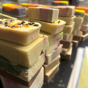 Puede incluir: Una pila de pastillas de jabón rectangulares en varios colores, incluyendo crema, verde, marrón y rosa. La pastilla superior presenta virutas de color naranja, negro y blanco. Algunas pastillas tienen patrones de colores en capas.