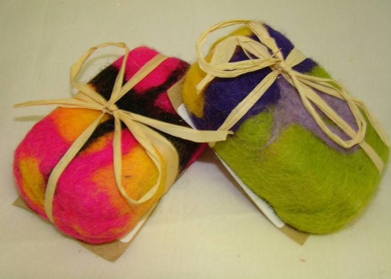 May include: Two handmade soap bars wrapped in natural twine. One bar is pink, yellow, and black, and the other is green, purple, and yellow.