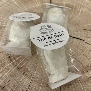May include: Two white packages containing bath tea, labelled "Thé de bain" for a better bath. The packages are made of clear plastic and white paper. The label also includes the text "Bain & Bulles Savonnerie Artisanale".