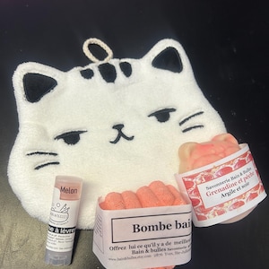 Op de afbeelding: Een witte katvormige washand met zwarte accenten, een lippenbalsem tube met het label "Melon", en twee pakketten badbruisballen. E&eacute;n badbruisbalpakket is gelabeld "Bombe bai", en de andere is gelabeld "Savonnerie Bain & Bulles Grenadine et p&ecirc;che Argile et soie".
