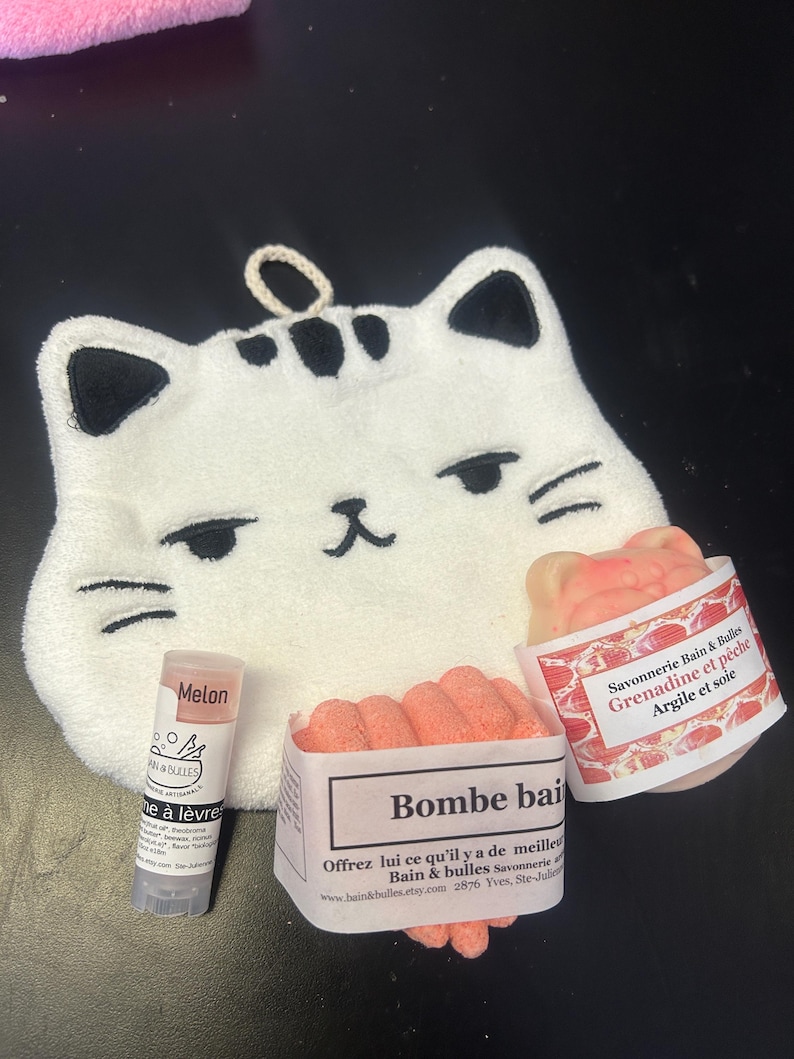 Op de afbeelding: Een witte katvormige washand met zwarte accenten, een lippenbalsem tube met het label "Melon", en twee pakketten badbruisballen. E&eacute;n badbruisbalpakket is gelabeld "Bombe bai", en de andere is gelabeld "Savonnerie Bain & Bulles Grenadine et p&ecirc;che Argile et soie".