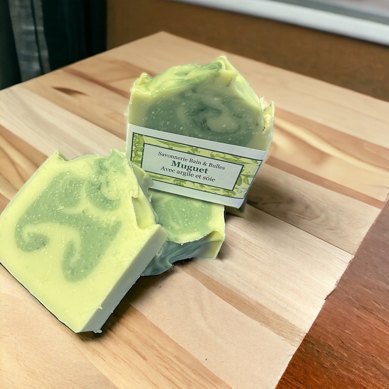 May include: Handmade soap bars with a layered design of green and yellow hues. The soap has a marbled appearance and is labeled with the text "Muguet Avec argile et soie". The soap is displayed on a wooden surface.