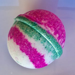 Puede incluir: Una bomba de baño esférica con una superficie texturizada que presenta rayas diagonales alternas en rosa intenso, blanco y verde bosque. El objeto tiene una costura distintiva alrededor del centro, creando un aspecto estratificado y granulado.