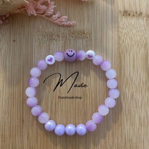 Könnte beinhalten: Lila-weißes Perlenarmband mit einem Smiley-Anhänger und Herz-Anhängern. Das Armband ist von "Mavie Handmadeshop" hergestellt.