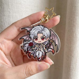 May include: A clear acrylic charm featuring a chibi vampire character with white hair, red eyes, and bat wings. The character wears a white and gold outfit and holds a rose and a staff. The charm has a gold star-shaped keychain attachment.