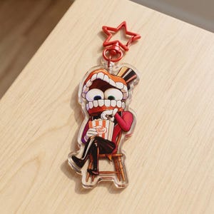 May include: A clear acrylic keyring featuring a cartoon character with a large mouth full of teeth, wearing a top hat and red suit, sitting on a wooden chair while eating popcorn from a striped bucket. The keyring has a red star-shaped clasp.
