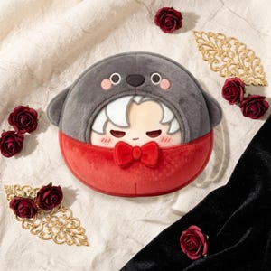 Puede incluir: Un peluche redondo con capucha gris con forma de foca y pajarita roja. La mitad inferior es roja y la cara del personaje es visible. Rosas rojas decorativas y detalles de filigrana dorada rodean el juguete.