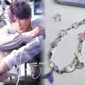May include: A beaded necklace with a mermaid theme. The necklace features clear, iridescent, and purple beads, along with seashell and starfish charms. A purple mermaid tail charm hangs at the end. A clear starfish is also visible.