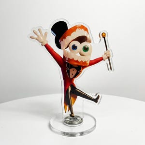 Puede incluir: Una figura de acrílico transparente que muestra un personaje de dibujos animados con sombrero de copa, frac rojo y cabeza con forma de dentadura con un ojo azul y otro verde. El personaje sostiene un bastón fino con una esfera dorada.