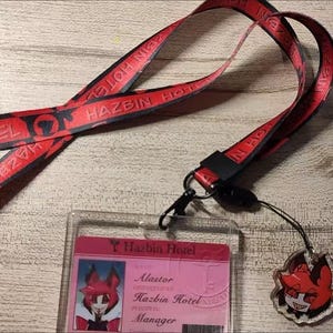 May include: A red lanyard with the text "HAZBIN HOTEL" in black. The lanyard is connected to a clear plastic ID holder with a pink card inside. A small charm with a cartoon character is attached.