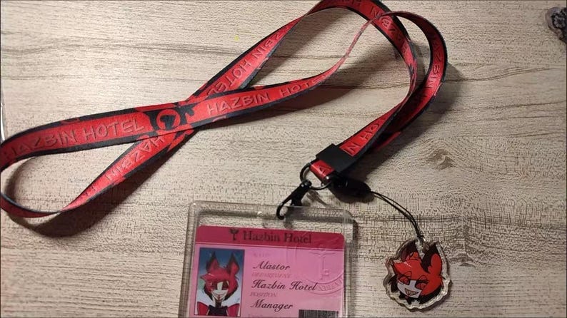 May include: A red lanyard with the text "HAZBIN HOTEL" in black. The lanyard is connected to a clear plastic ID holder with a pink card inside. A small charm with a cartoon character is attached.