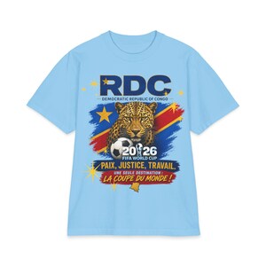 May include: Light blue t-shirt featuring a graphic of a leopard, a soccer ball, and the text "RDC - DEMOCRATIC REPUBLIC OF CONGO" and "2026 FIFA WORLD CUP". The shirt also includes the phrases "PAIX, JUSTICE, TRAVAIL" and "LA COUPE DU MONDE!"