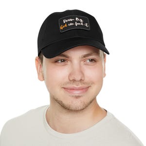May include: Black baseball cap with a rectangular patch featuring the text "Dream Big, God can fund it" in a mix of orange, yellow, and white. The cap is worn by a person.