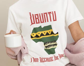 May include: White t-shirt with the word "UBUNTU" in red, above a green and yellow map of Africa. The phrase "I AM BECAUSE WE ARE" is printed below the map in red. The shirt is made of a soft material.