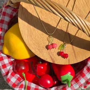 May include: Two gold necklaces with cherry charms. The necklaces are displayed in a wicker basket with red and white checkered lining. The basket also contains a lemon and red cherries.