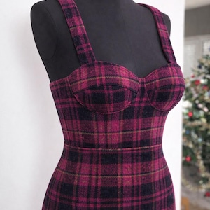 May include: A bustier-style dress in a plaid pattern, displayed on a black mannequin. The dress features a fitted bodice with structured cups and thin shoulder straps. The fabric is a mix of burgundy, black, and gold tones.