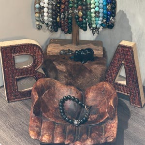 Könnte beinhalten: Eine Präsentation von Armbändern mit Perlen in verschiedenen Farben, darunter Schwarz, Grün und Blau, auf einem Holzständer. Ein schwarzes Perlenarmband mit einem silbernen Anhänger liegt in einer geschnitzten Holzhand. Dekorative Holzbuchstaben mit der Aufschrift "BRA".