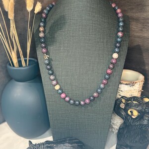 Puede incluir: Un collar de cuentas con una mezcla de piedras azul oscuro, rosa y crema se exhibe en un maniquí gris. Un pequeño dije plateado está adjunto. El collar es parte de una colección de joyas.