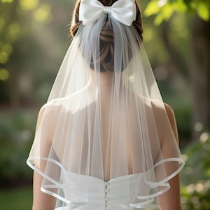 Puede incluir: Un velo de novia blanco con un lazo de satén y ribete de cinta. El velo está sujeto a la parte posterior de un vestido de novia blanco. La imagen está tomada desde atrás, con un fondo verde borroso.