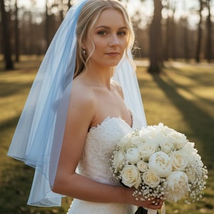 May include: A bride in a strapless white lace wedding dress holds a bouquet of white roses and baby's breath. She wears a light blue veil. The background is a sunlit park with trees.