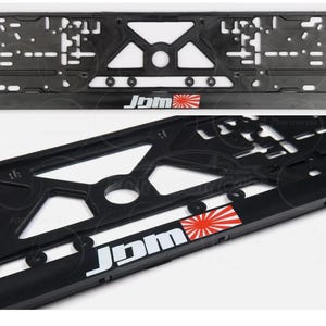 May include: Black license plate frame with a geometric design. The frame features the text "JDM" in white, with a red and white rising sun emblem. The frame is designed to hold a license plate.