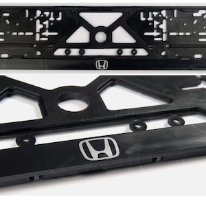 Può includere: Cornice nera per targa con logo Honda argentato. La cornice presenta un design geometrico complesso con molteplici aperture e fori di montaggio. Realizzata in materiale resistente, è progettata per una targa standard.