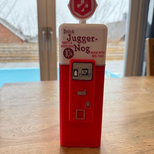 Peut inclure: Distributeur de carburant de style vintage rouge et blanc avec les mots "Drink Jugger-Nog That's a good nog 10¢ Made with real eggs" sur le devant.