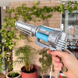 Peut inclure: Un jouet pistolet argenté et bleu avec un flocon de neige sur le côté. Le pistolet a un long canon et une poignée. Le pistolet est tenu par la main d'une personne.