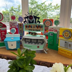 Pode incluir: Uma coleção de máquinas de venda automática coloridas e estilizadas com vários designs e texto, incluindo "Pack-a-Punch" e "Double Tap II." As máquinas estão dispostas sobre uma superfície de madeira, com uma planta verde em primeiro plano.